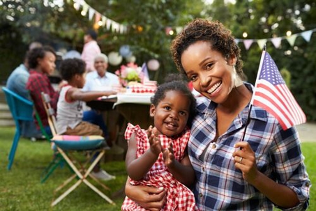 Young African American mother holding a baby girl in one hand and an American flag in the other in front of 4th of July celebration