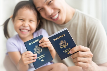 A grinning immigrant father with his little girl holding up their passports to the camera.
