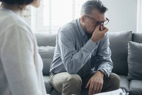 A husband tiredly kneads the bridge of his nose as his wife stands to confront him during divorce.