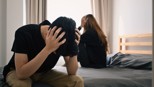 Two fighting spouses not facing each other sitting on opposite sides of the bed.