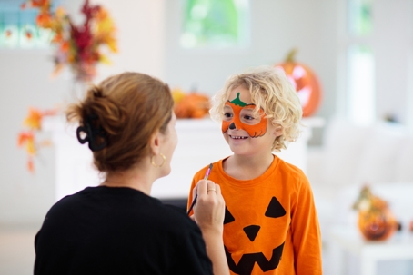 a mother paints a pumpkin on her child's face while preparing for a costume during Halloween