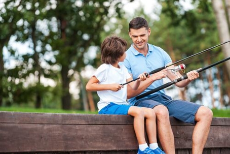 Dad teaching son to fish on dock in forest. Both wearing blue. Photo represents paternity.
