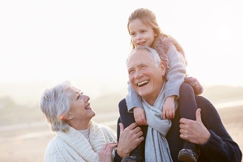 Smiling grandpa carries his granddaughter on his shoulders while her smiling grandma looks on.