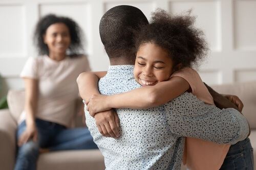 Smiling girl hugs her father while her sitting mother smiles in the background.